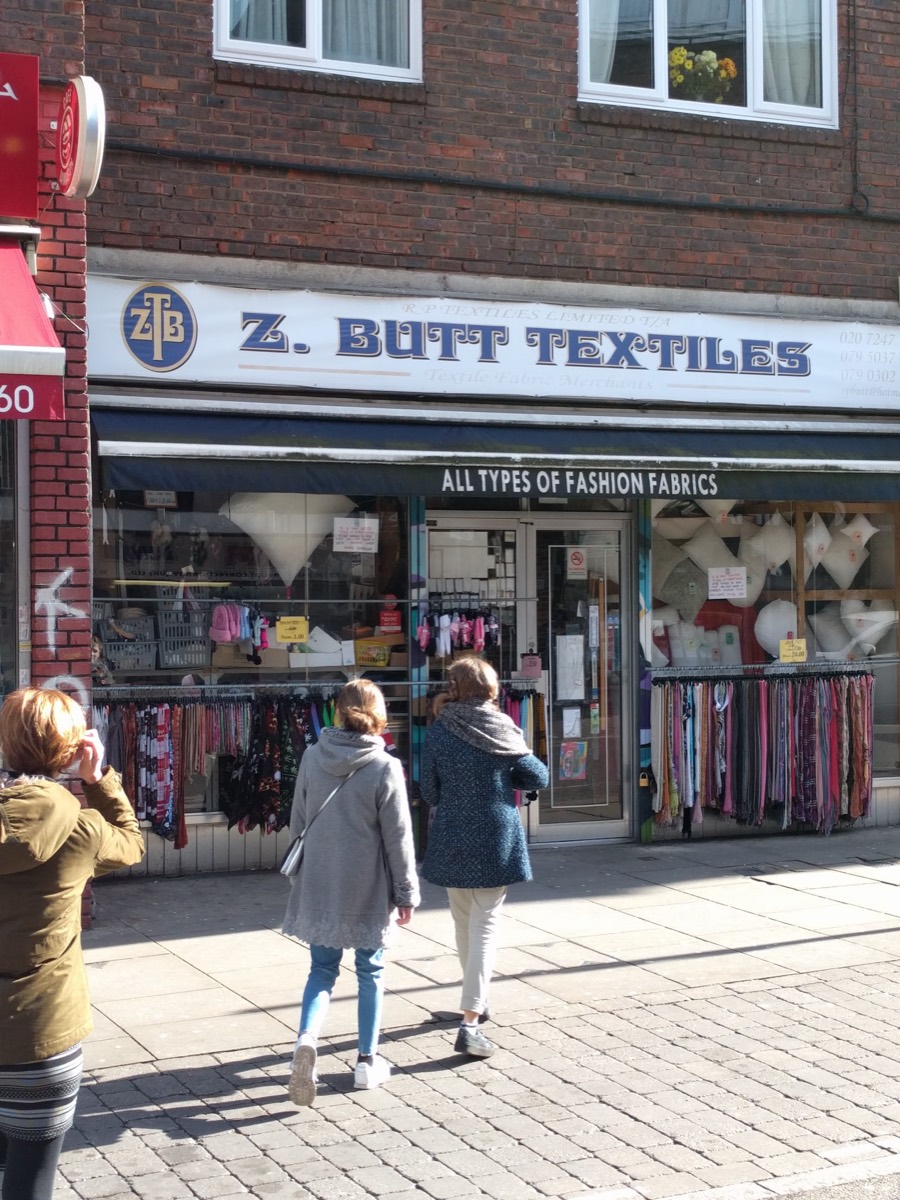 The navy and gold shopfront of Z. Butt Textiles on Brick Lane, with rails of fabric outside and shoppers passing in the sun.